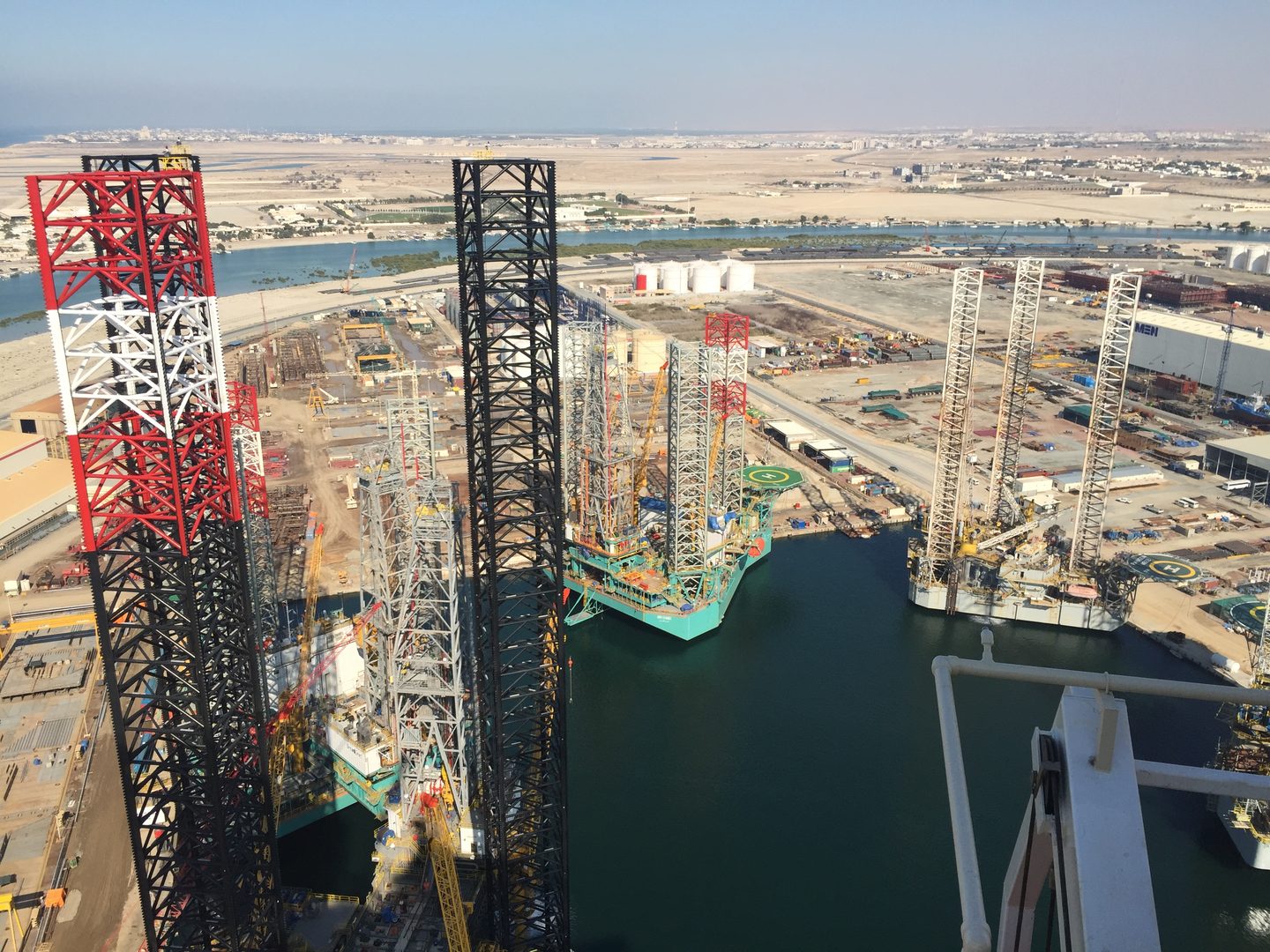 Elevated view from a jack-up during jacking trials, overlooking rigs and shipyard activity with lifeboat-davit and winch equipment in the foreground